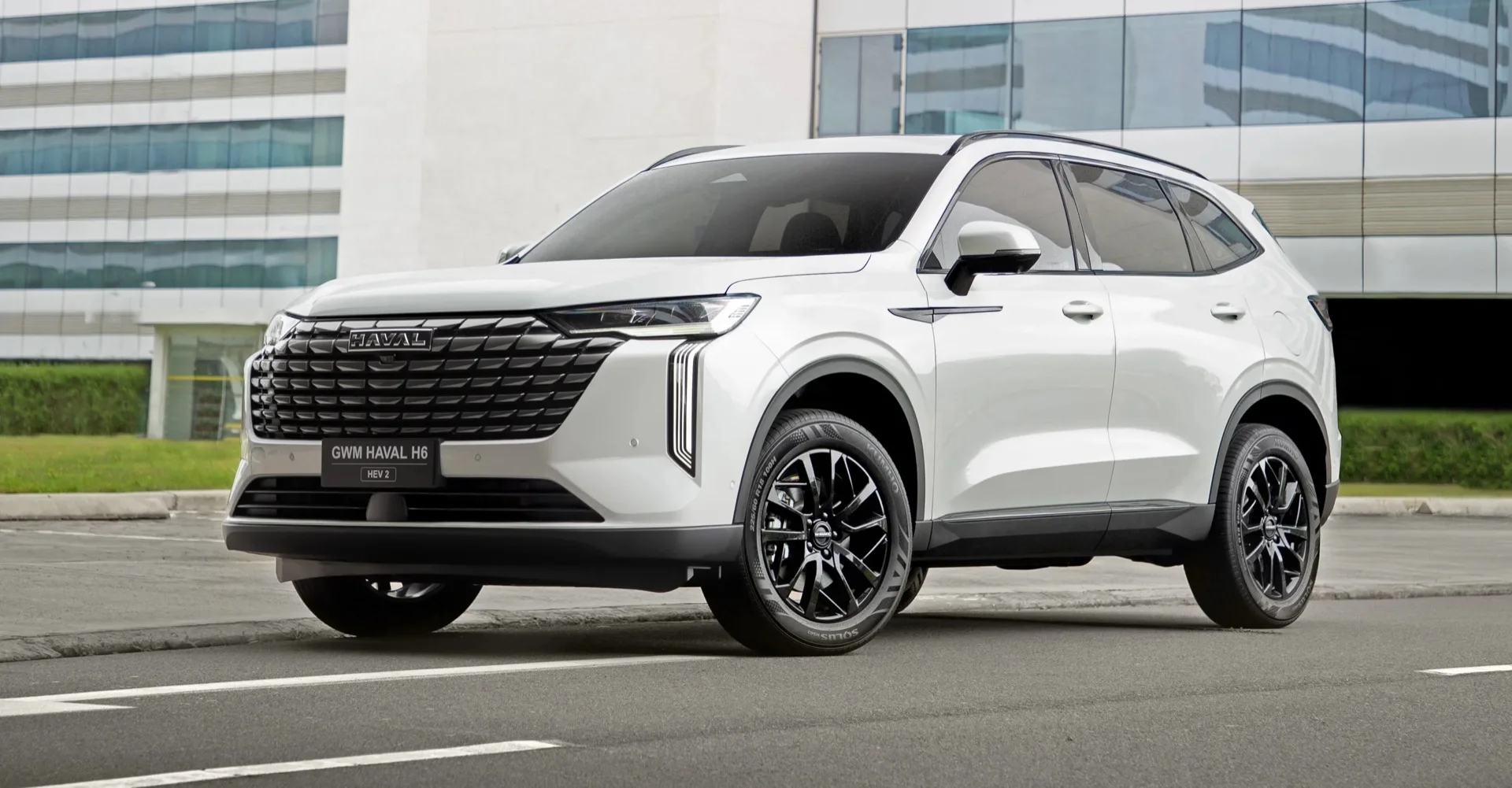 GWM Haval H6 HEV2 branco com rodas pretas, visto de frente em ângulo de três quartos diante de um prédio envidraçado