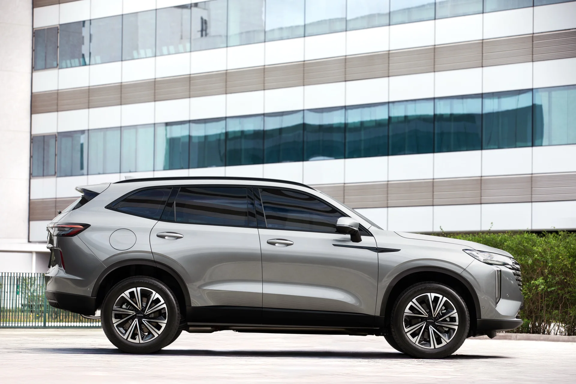 Lateral do GWM Haval H6 PHEV19 prata com rodas aro 19, estacionado em frente a um prédio de vidro
