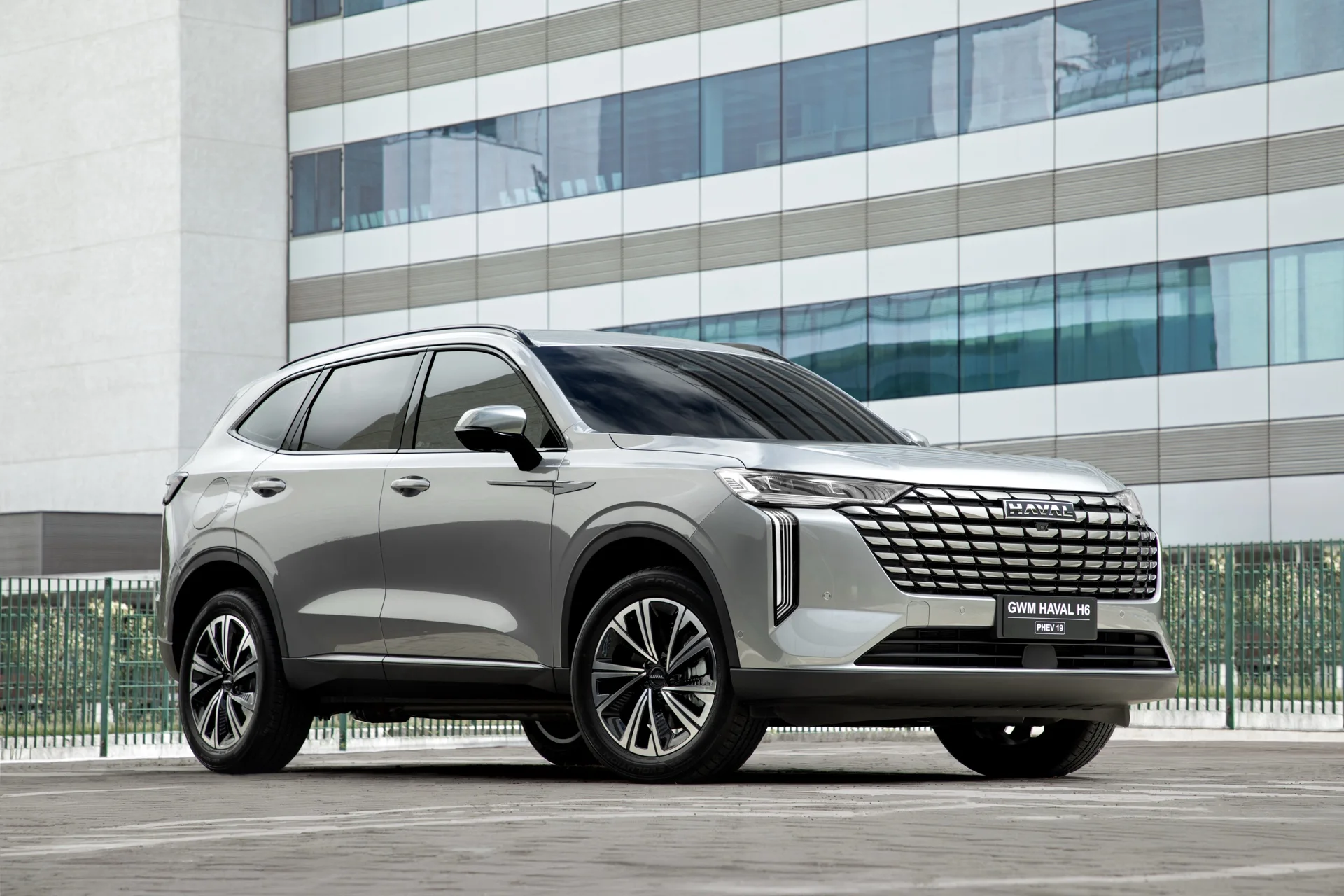 GWM Haval H6 PHEV19 prata visto de frente, em ângulo de três quartos, diante de um prédio envidraçado