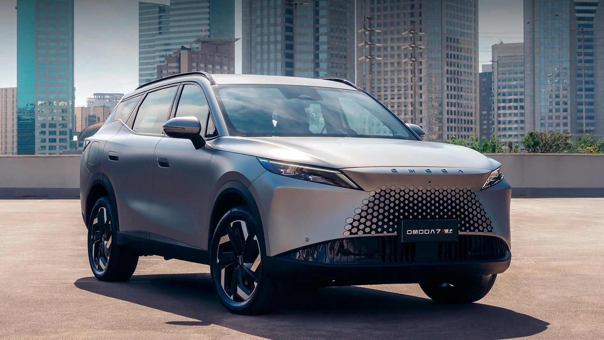 Omoda 7 SHS-P cinza visto de frente, de três quartos, estacionado em um terraço com prédios ao fundo